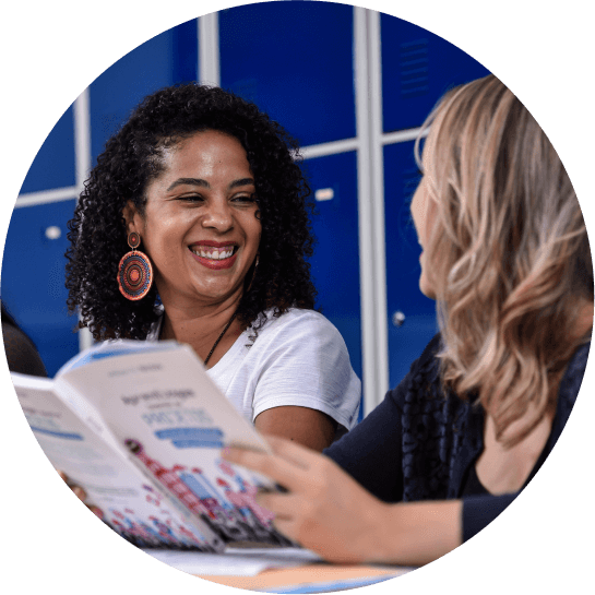 Duas mulheres sorriem se olhando com um livro na mão em uma mesa